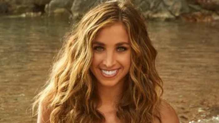 Katie Austin poses in front of the water in a bikini and wears her dirty blonde hair in a soft wave.